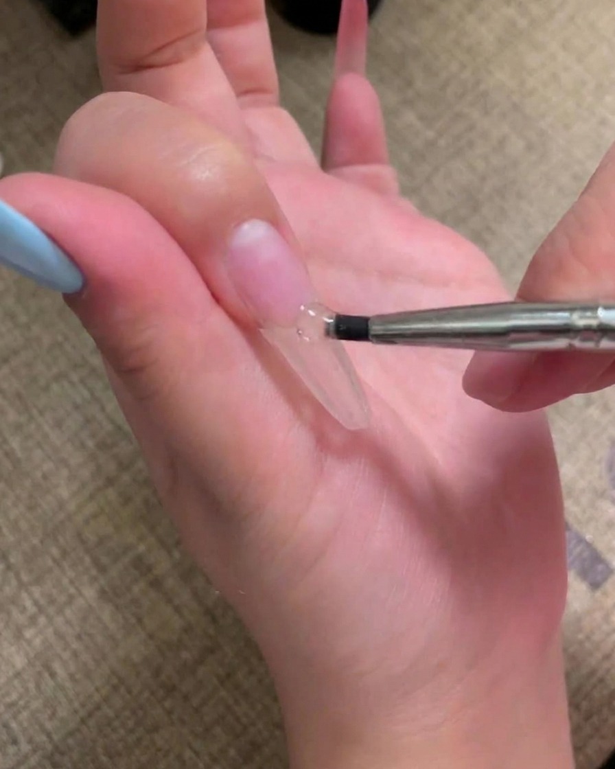 Un pinceau appliquant une couche de gel rose chair sur un ongle préparé.
