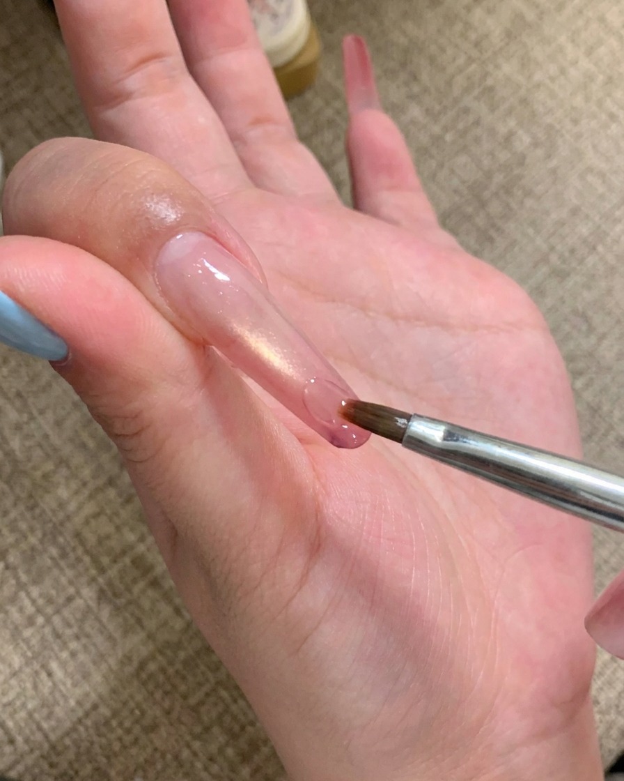 Un pinceau nail art appliquant une goutte de gel adhésif transparent sur le bout dun ongle long.
