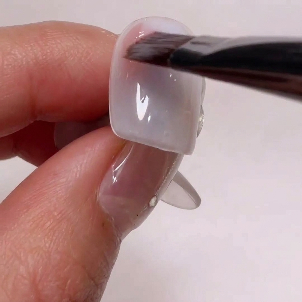 Un pinceau de nail art appliquant un gel rose nude sur les coins dune capsule.