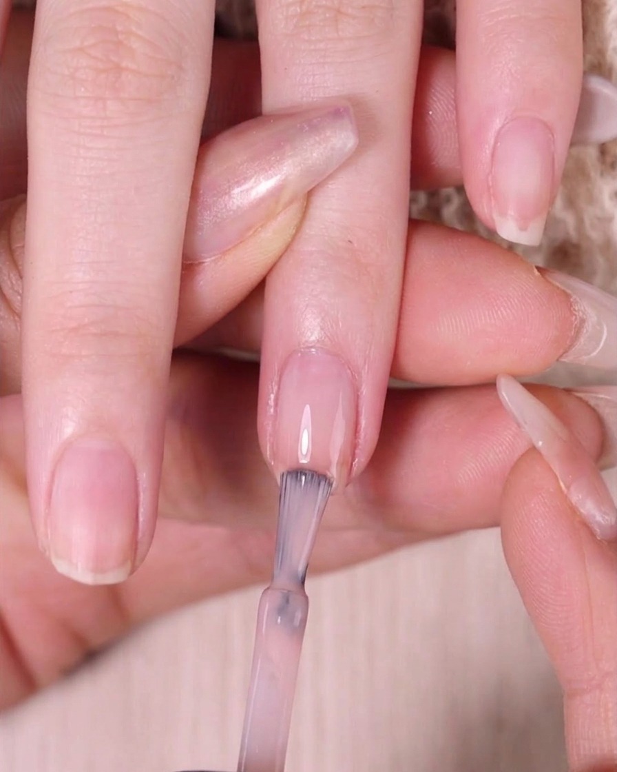 Un pinceau à ongles appliquant du vernis rose nude transparent uniquement sur le tiers supérieur de longle.