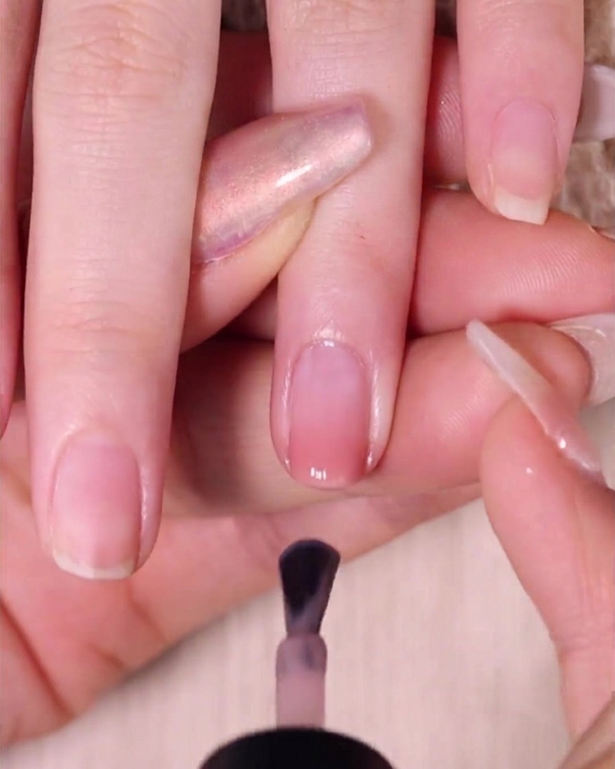 Un pinceau à vernis appliquant une couche lisse de vernis rose nude transparent sur un ongle ombré, en laissant un petit espace près de la cuticule.