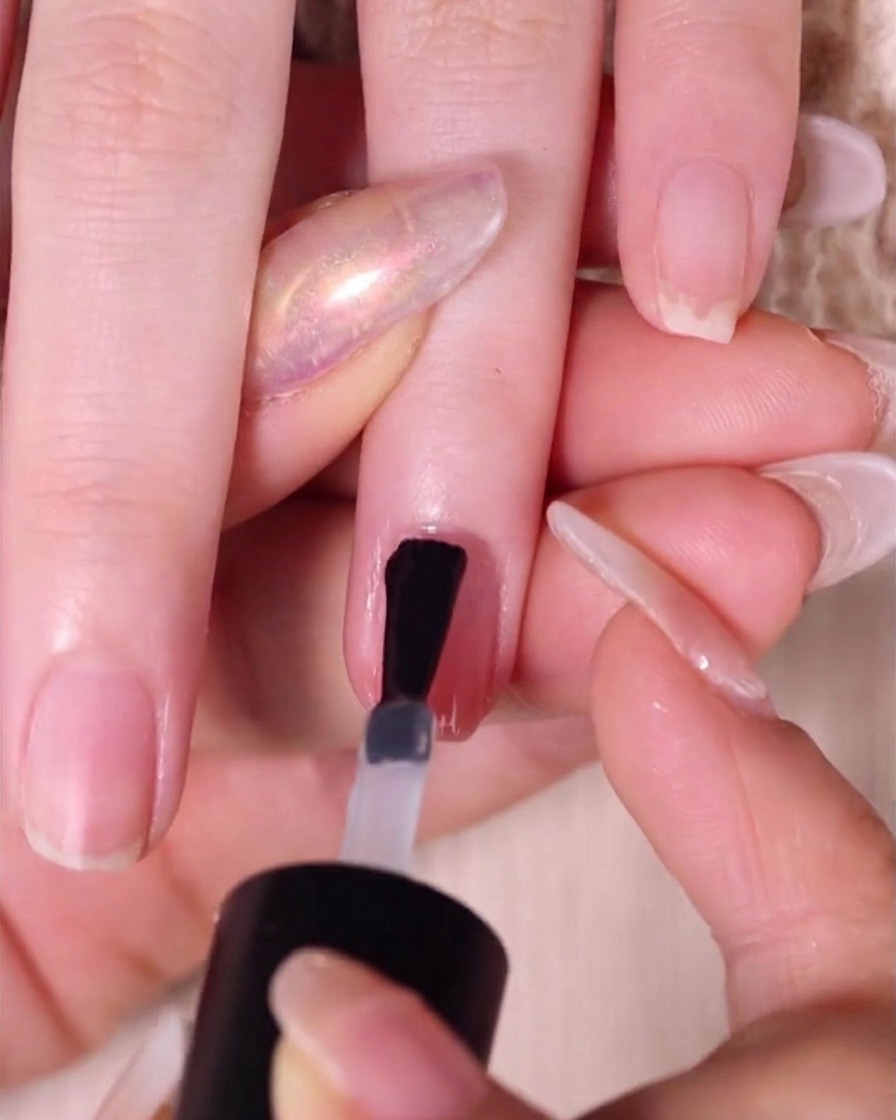 Un pinceau appliquant une couche épaisse et brillante de top coat transparent sur un ongle ombré rose nude terminé.