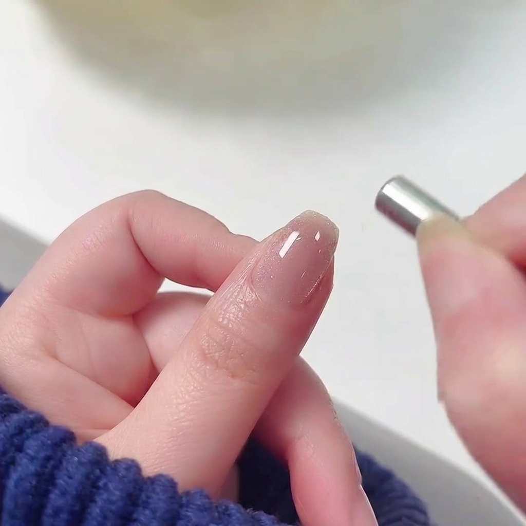 Un petit aimant cylindrique tenu près de la cuticule dun ongle recouvert de vernis gel cat-eye rose humide et scintillant.