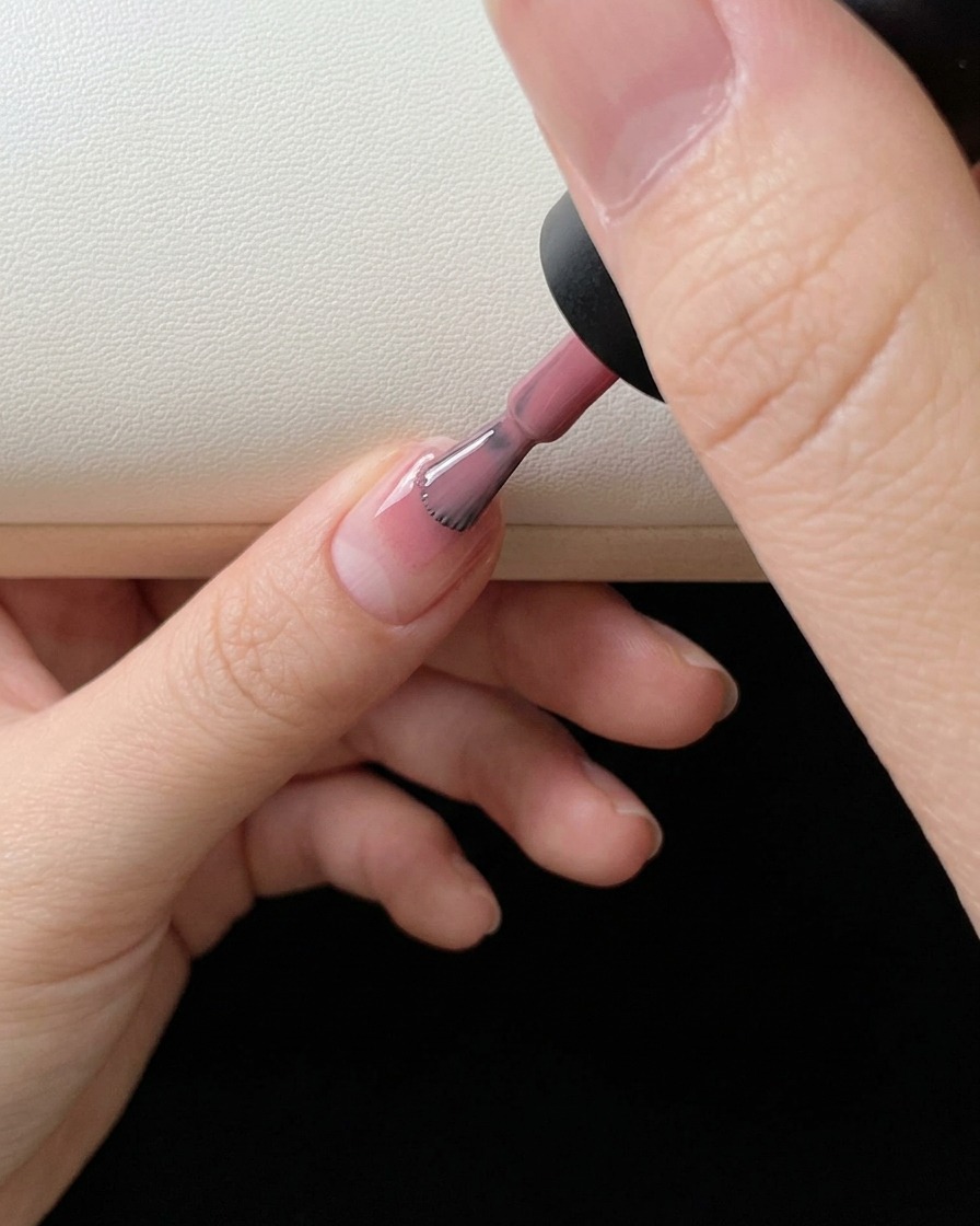 Gros plan sur un pinceau à vernis appliquant du gel rose sur le bout de longle naturel.