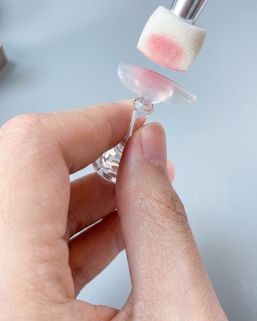 Gros plan dune éponge appliquant une douce couleur rose poudré sur une capsule posée sur un support.