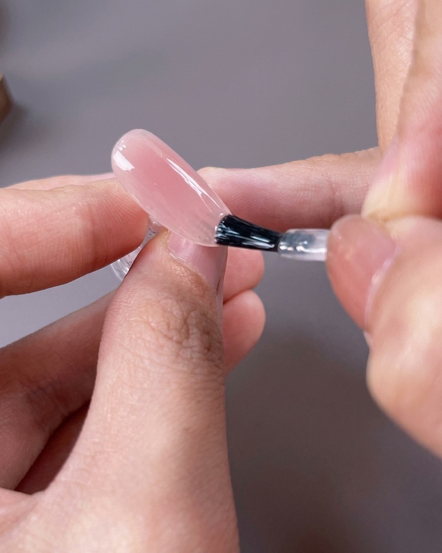 Pinceau appliquant un vernis gel nude rosé clair et translucide sur toute la surface de longle.