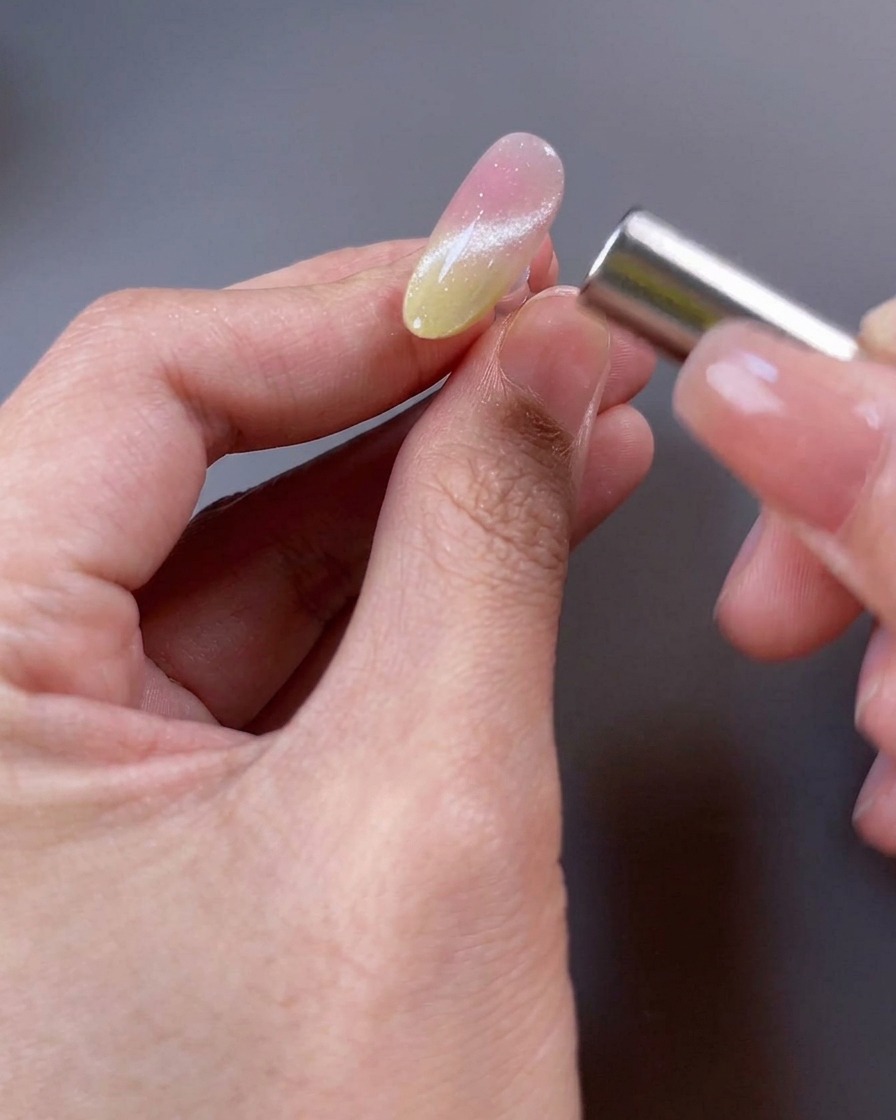 Gros plan dun ongle avec du vernis gel magnétique, où un aimant est tenu près de la surface pour manipuler les particules et former un large faisceau lumineux.