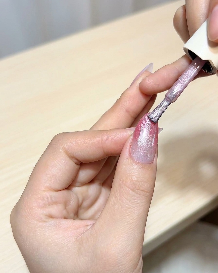 Un pinceau appliquant une couche de vernis gel œil de chat sur une base ombrée magenta.