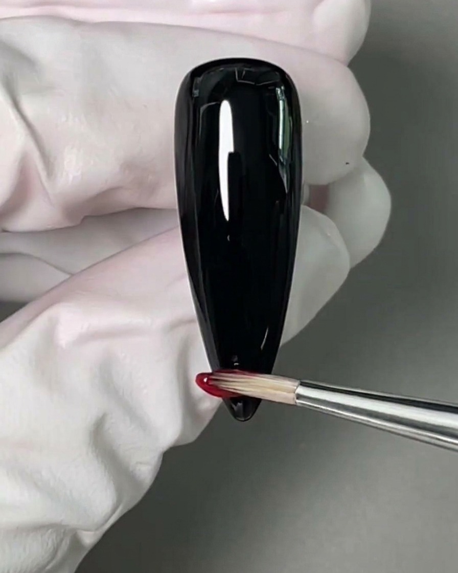 Un pinceau fin appliquant un vernis gel rouge vif sur le bout de longle noir.