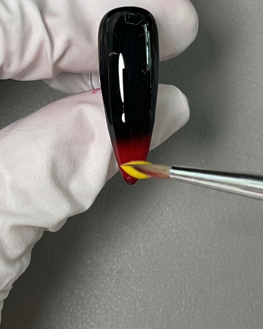 Un pinceau de précision déposant une goutte de vernis jaune juste au-dessus du rouge humide.