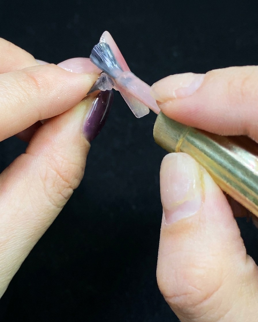 Un pinceau appliquant une fine couche de vernis nude sur une capsule transparente.