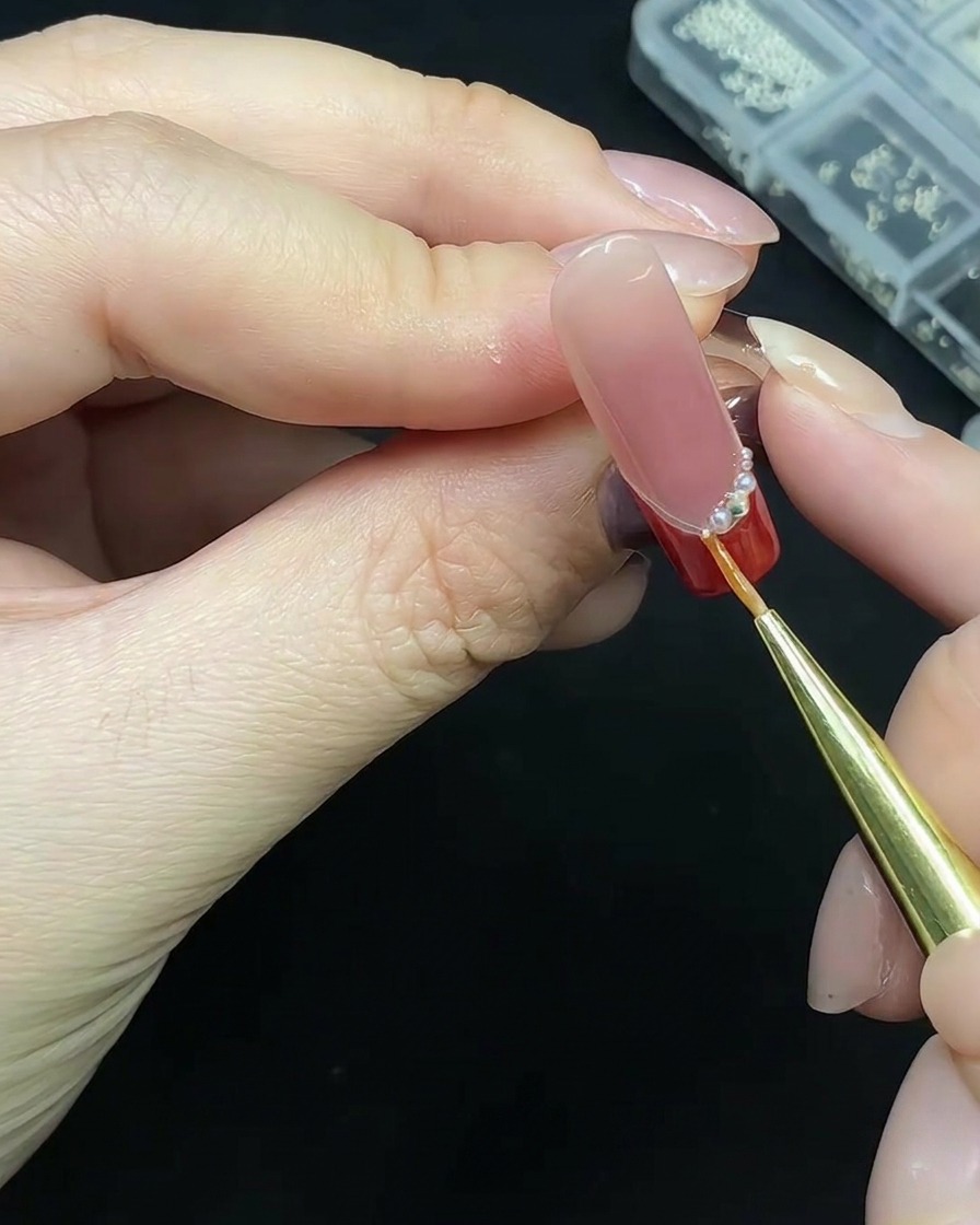 Un outil de précision plaçant des petites perles le long de la base dune French manucure rouge.