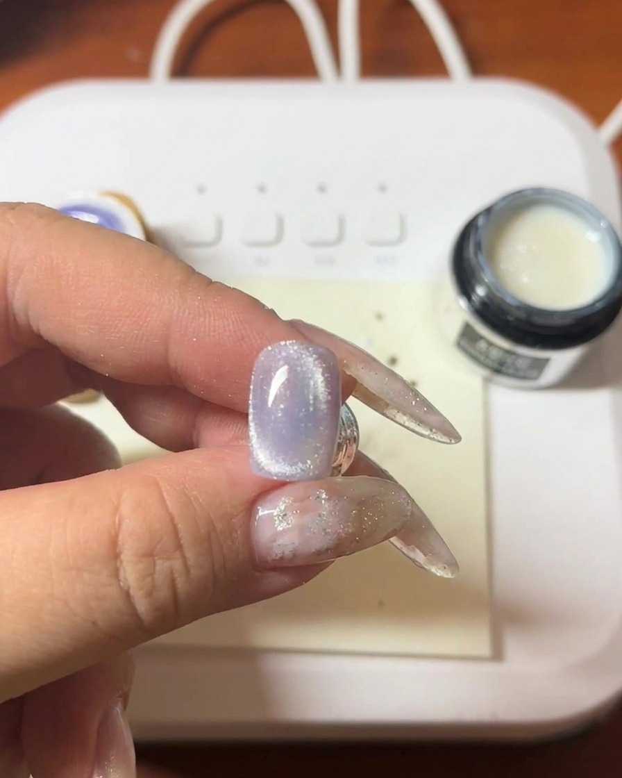 Gros plan dune main tenant une baguette magnétique au-dessus dun ongle en gel violet, montrant la traînée lumineuse se former.