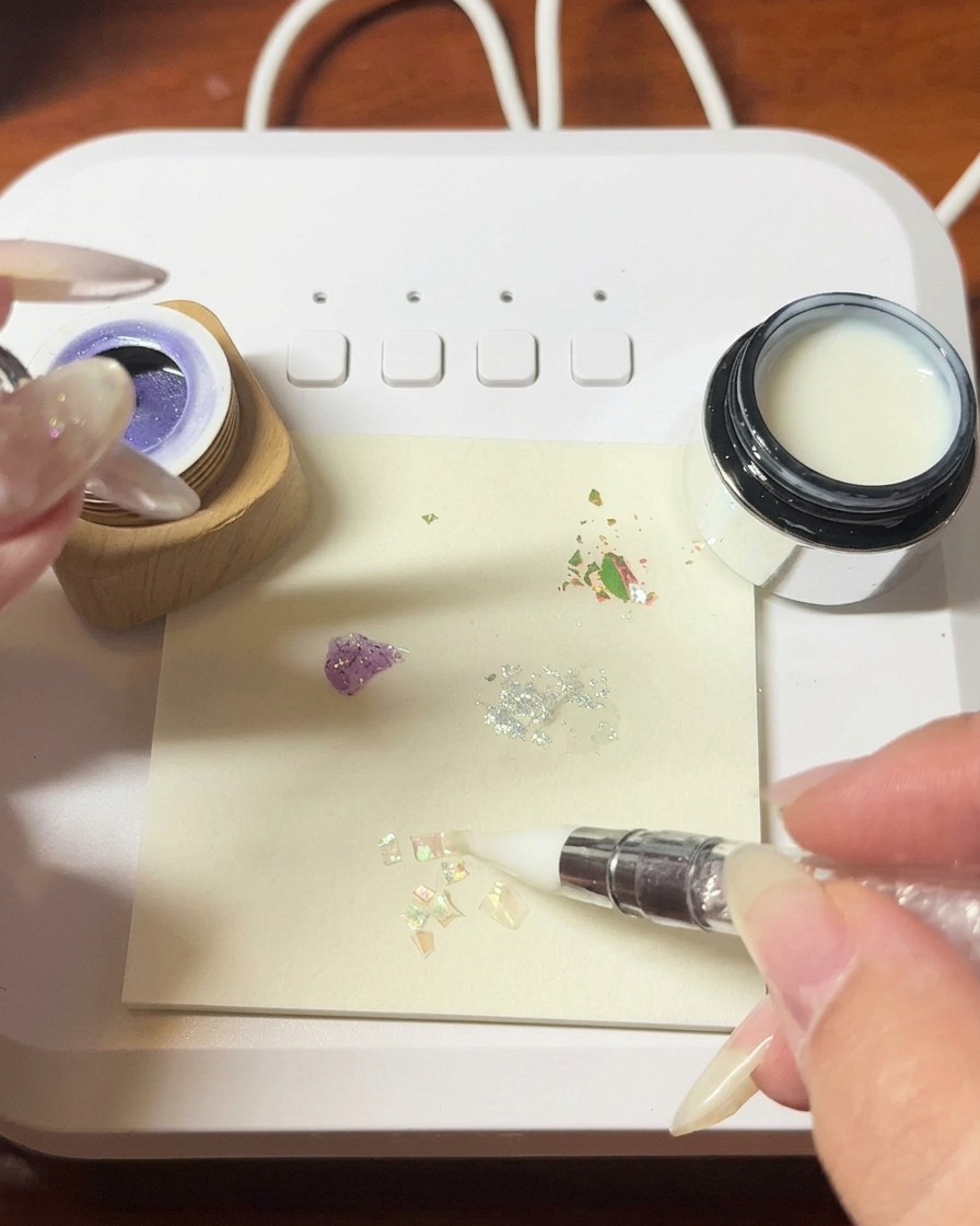 Une pince à épiler plaçant des fragments de coquillages irisés délicats sur une surface de gel humide.