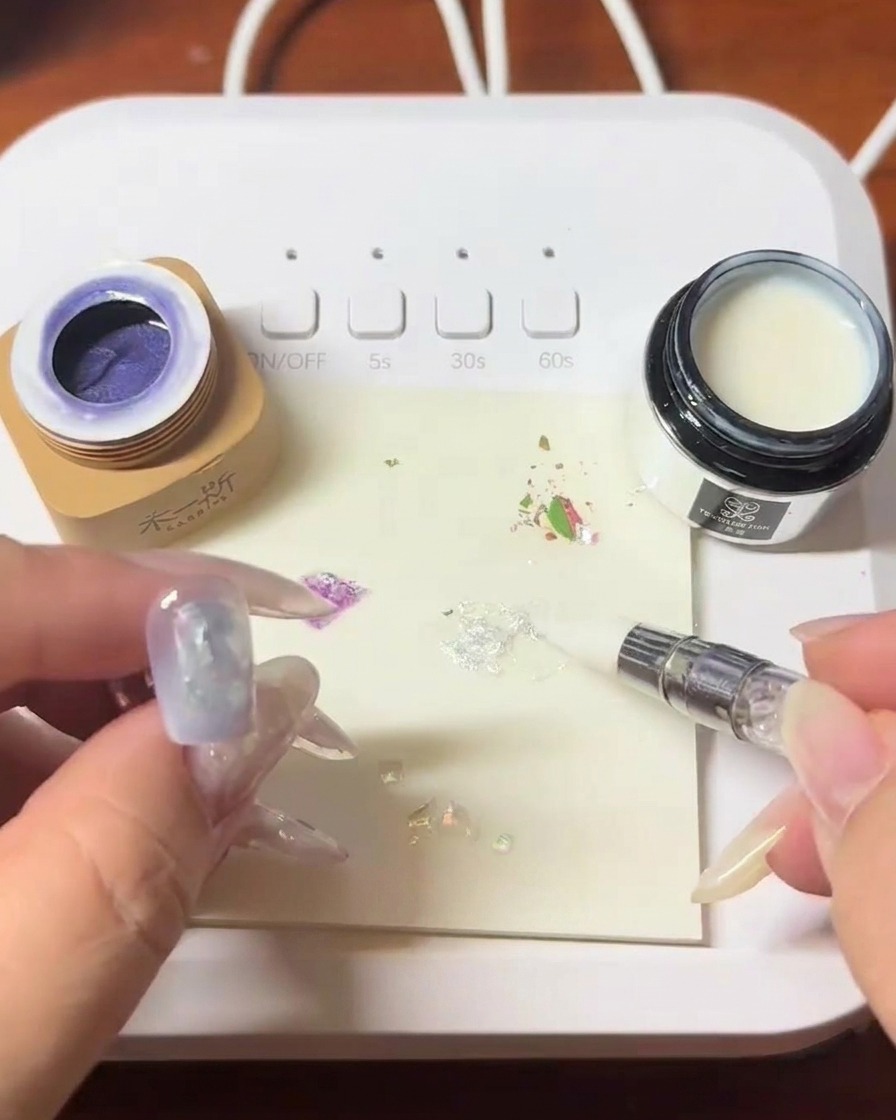 Un pinceau appliquant un blooming gel transparent sur longle décoré de coquillages.