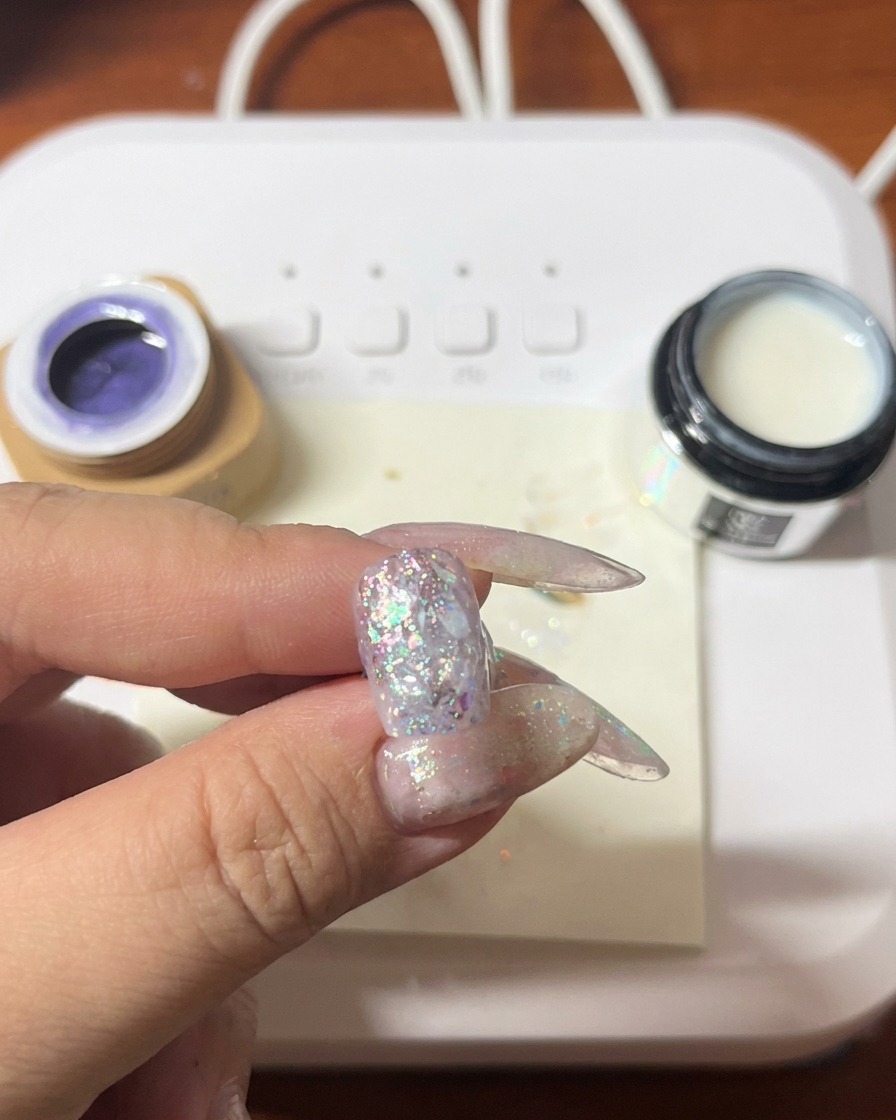 Un pinceau appliquant du vernis gel violet sur un ongle déjà décoré de paillettes et de coquillages.