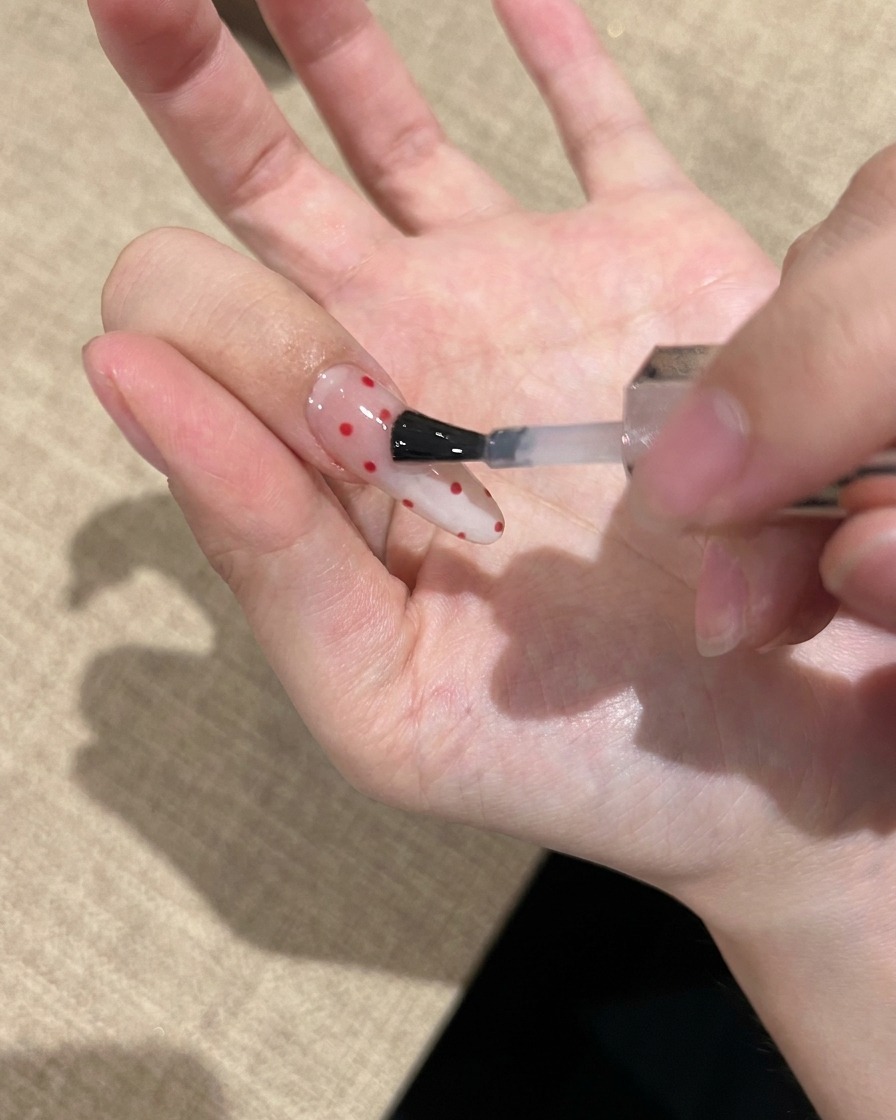 Gros plan dun pinceau à vernis appliquant un top coat transparent et brillant sur un ongle décoré de pois rouges.