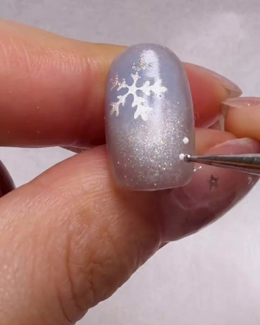 Vue rapprochée dun outil de pointage appliquant de minuscules points blancs et bleu clair autour dun flocon argenté sur un ongle bleu scintillant.