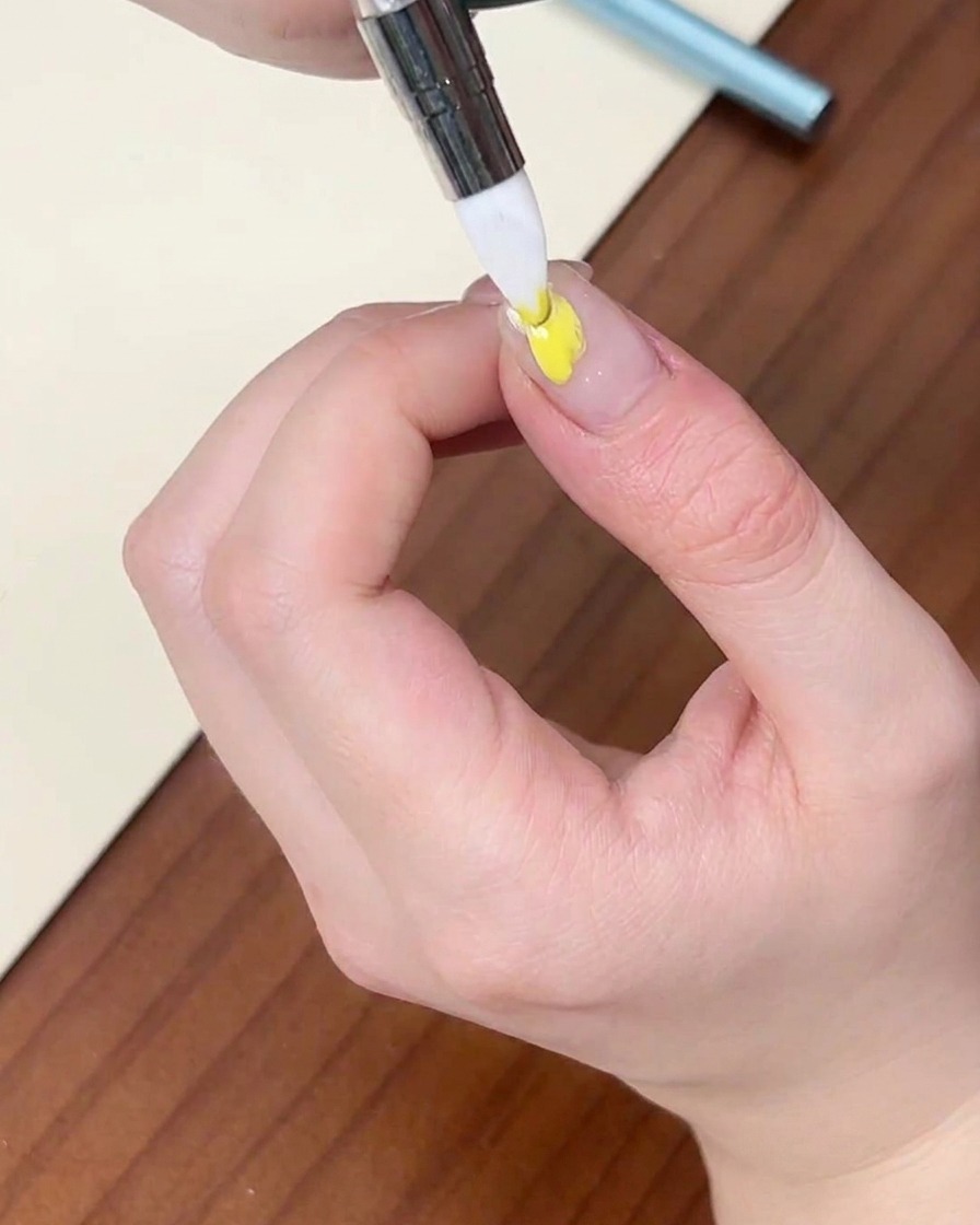 Un pinceau liner fin appliquant un point de vernis gel jaune sur un ongle naturel.