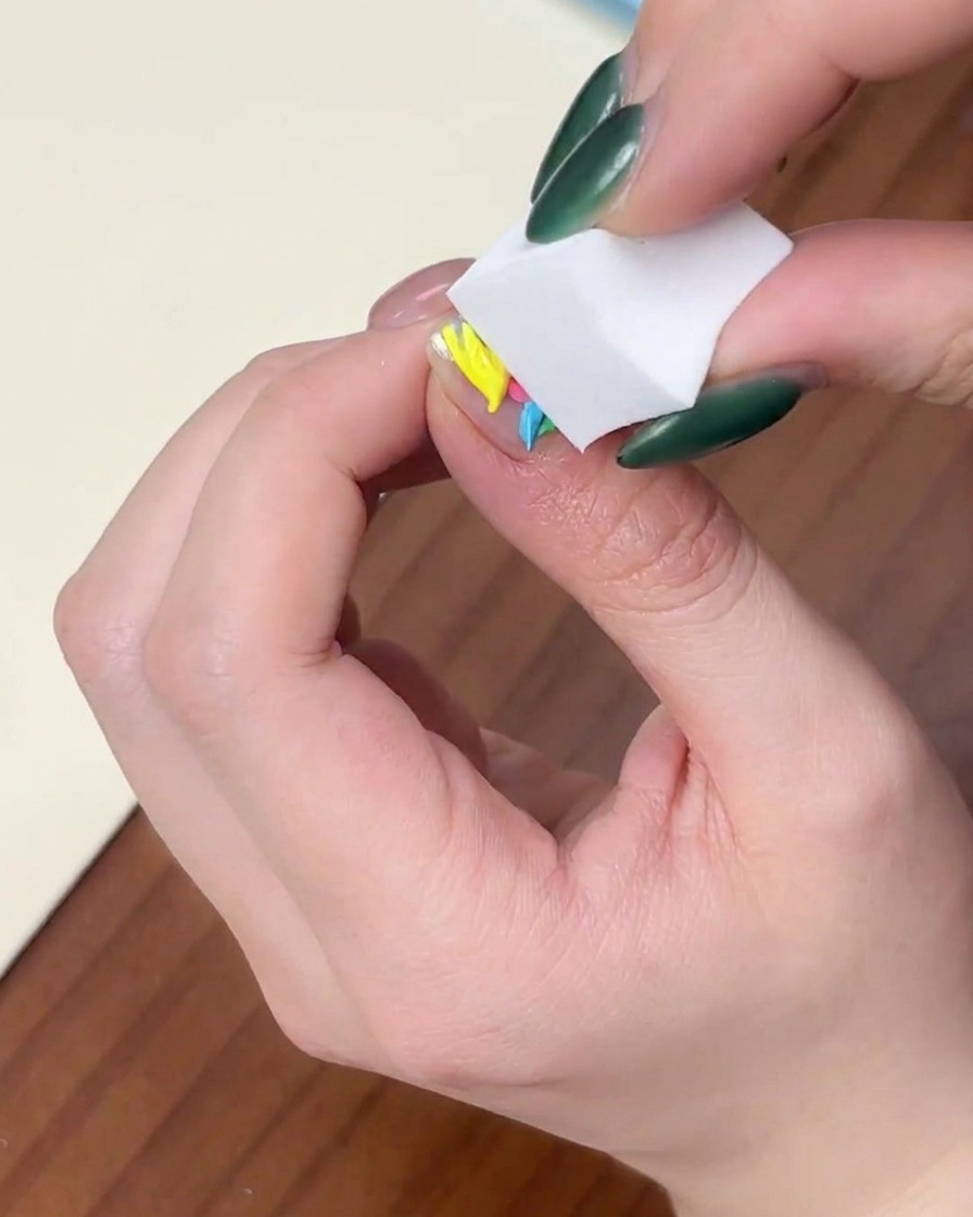 Gros plan dune main pressant une éponge à maquillage blanche verticalement sur un ongle couvert de points de vernis gel colorés.