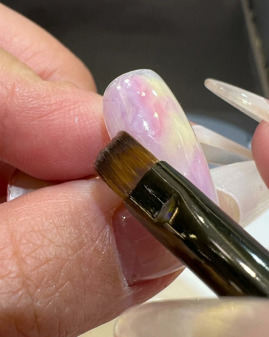 Un pinceau plat appliquant une couleur gel rose-violet diaphane sur le coin dun tips dongle à base blanche.