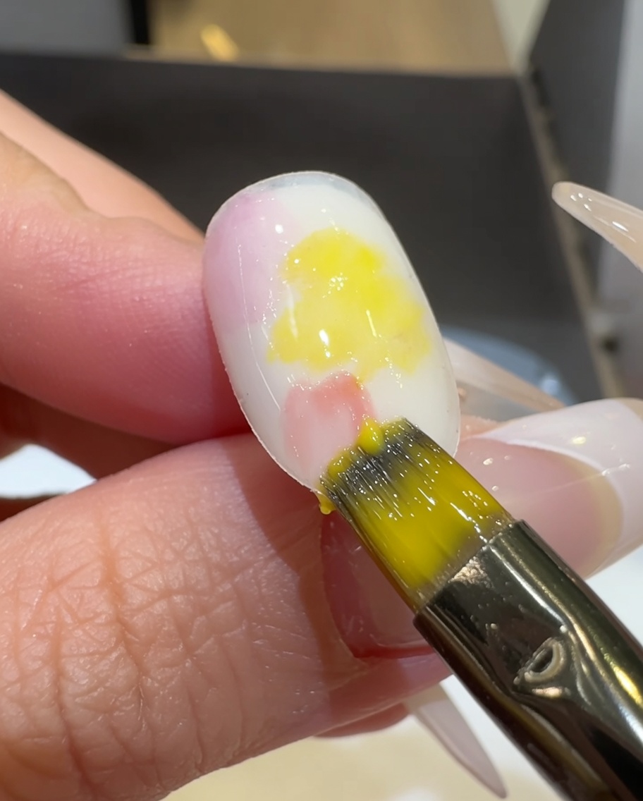 Un pinceau plat mélangeant le vernis gel rose et jaune sur la surface de longle.
