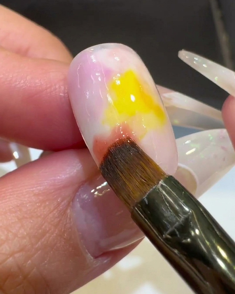 Gros plan sur un pinceau à ongles mélangeant le vernis gel rose dans le centre jaune, créant un effet dégradé.