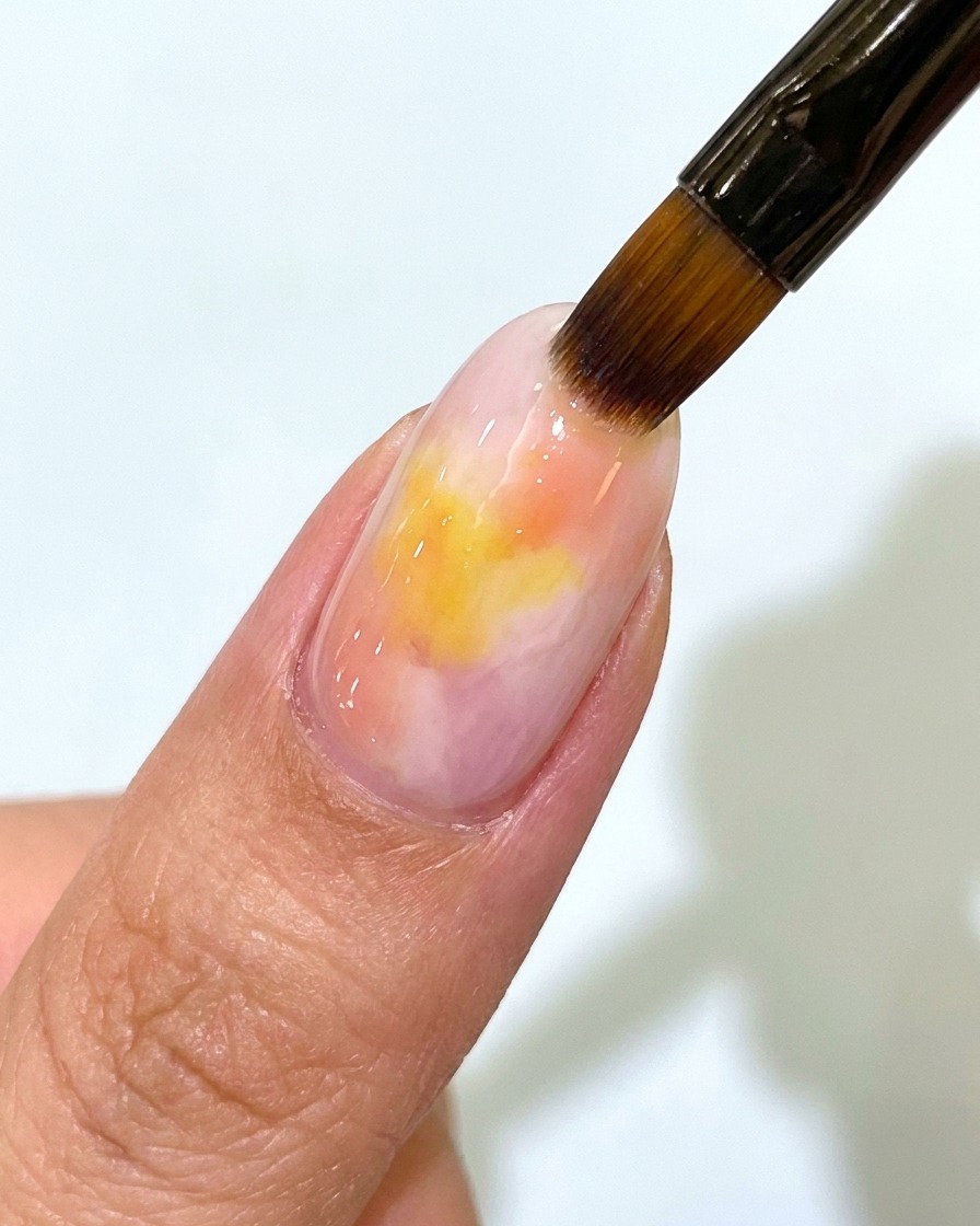 Un pinceau à ongles appliquant une couche de finition mate transparente sur un ongle en gel dégradé coucher de soleil rose et jaune.