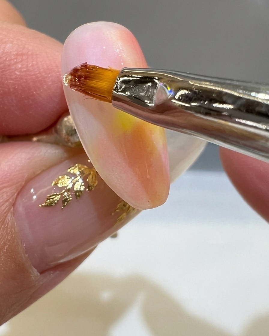 Un pinceau liner fin appliquant une ligne épaisse de gel 3D transparent sur un ongle mat couleur coucher de soleil.