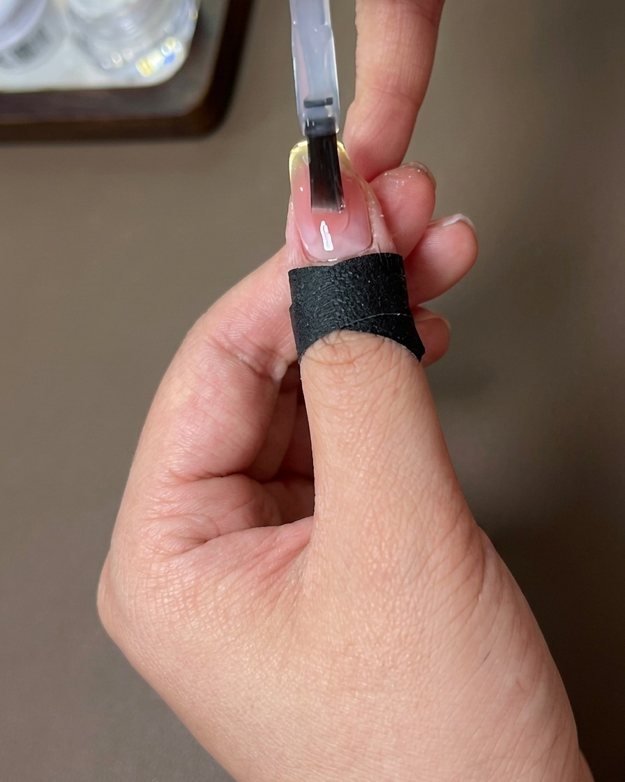 Un pinceau appliquant un top coat transparent brillant sur un ongle avec un dégradé sunset-blush et une French magnétique.