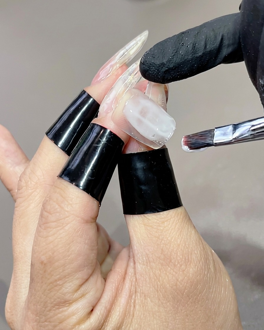 Une main avec de longues capsules transparentes sur laquelle un pinceau plat applique une base en gel blanc laiteux sur lannulaire.