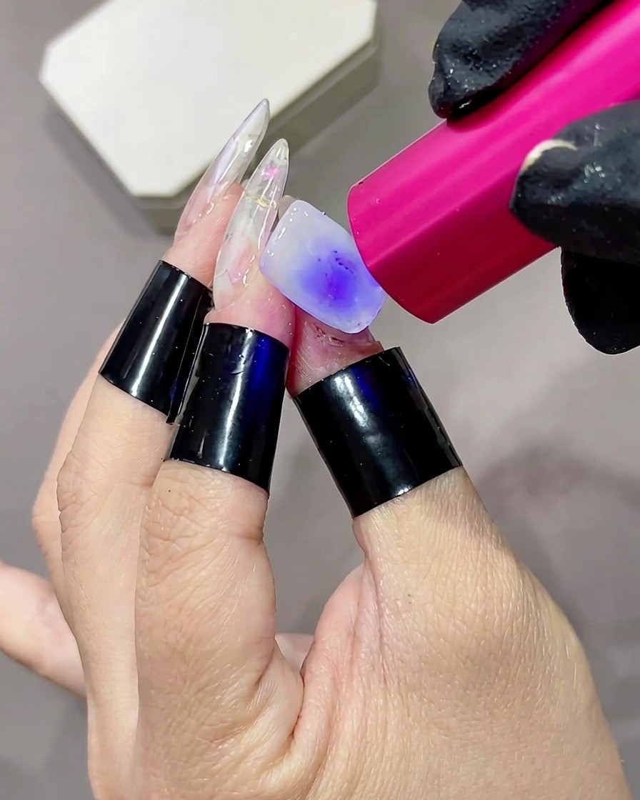 Gros plan dun doigt avec une base blanc laiteux en train dêtre catalysée par une petite lampe torche UV/LED rose.