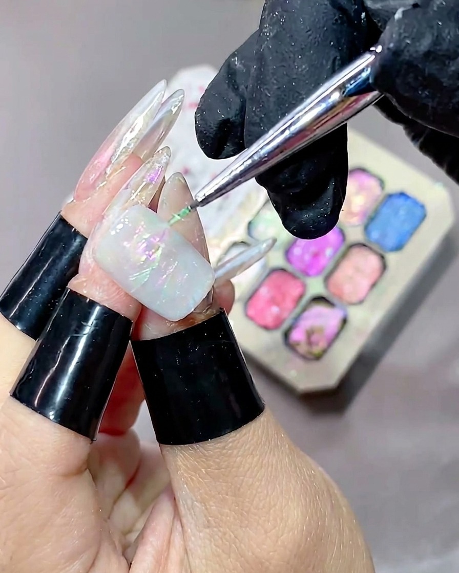 Un pinceau fin appliquant du gel texturé vert sur longle, avec une palette de gels colorés visible en arrière-plan flouté.
