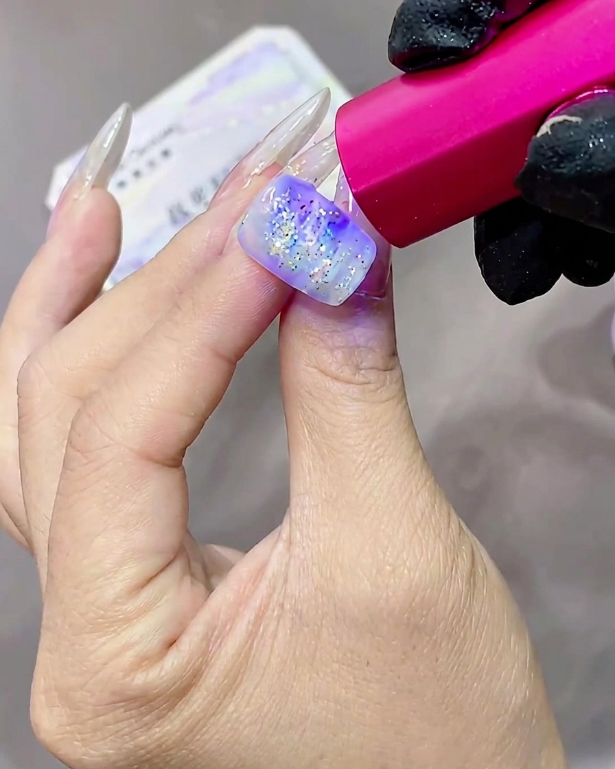 Une main avec des ongles en gel pailletés et texturés en train de catalyser sous une lampe UV teintée de rose.