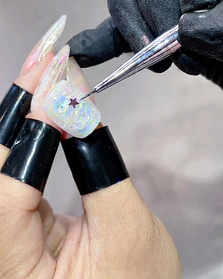 Un outil en métal plaçant une petite étoile noire au centre dun ongle recouvert de gel iridescent et multicolore.