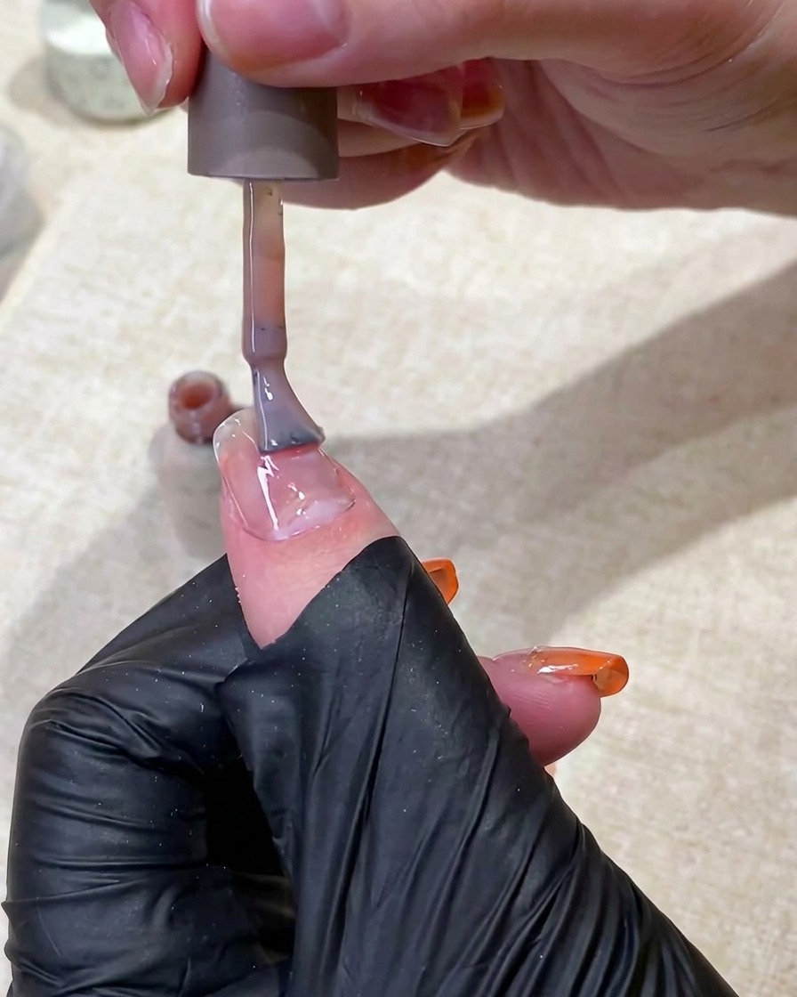 Une main gantée de noir utilisant un pinceau pour appliquer une base en gel nude translucide sur un ongle.