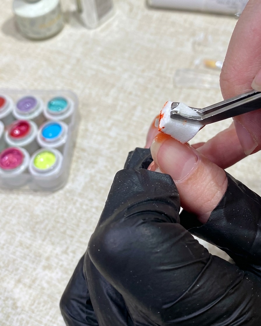 Une main gantée utilisant une pince pour tapoter du gel orange vif avec une petite éponge sur la pointe dun ongle.