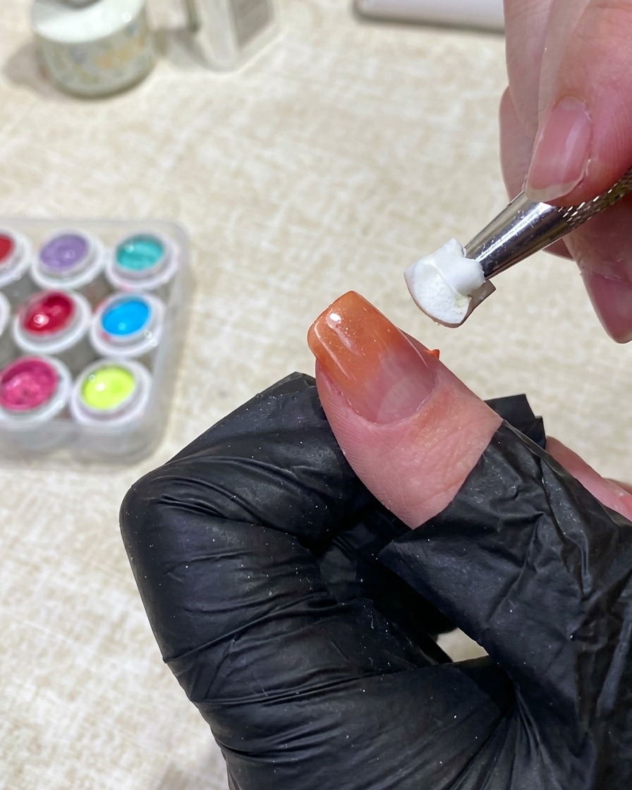 Un outil tenant un morceau déponge blanche et propre, tapotant doucement le milieu dun ongle pour diffuser le gel orange dans la base nude.