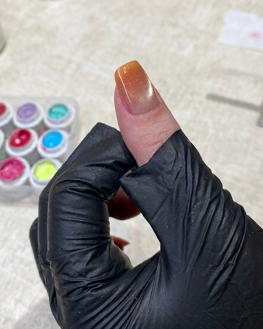 Une main gantée de noir utilise une pince pour tapoter doucement du gel orange vif avec une éponge sur la pointe de longle.
