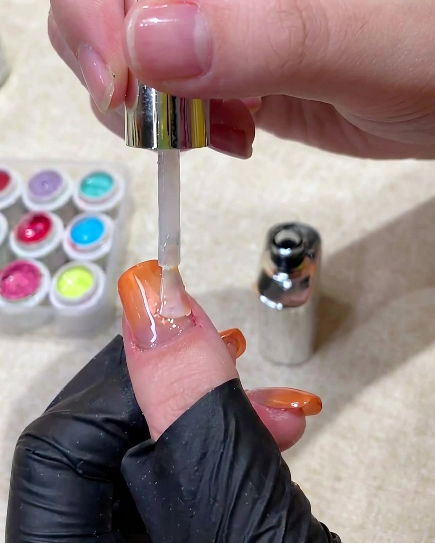 Un pinceau appliquant un top coat transparent et brillant sur un ongle terminé en dégradé orange et cat-eye.