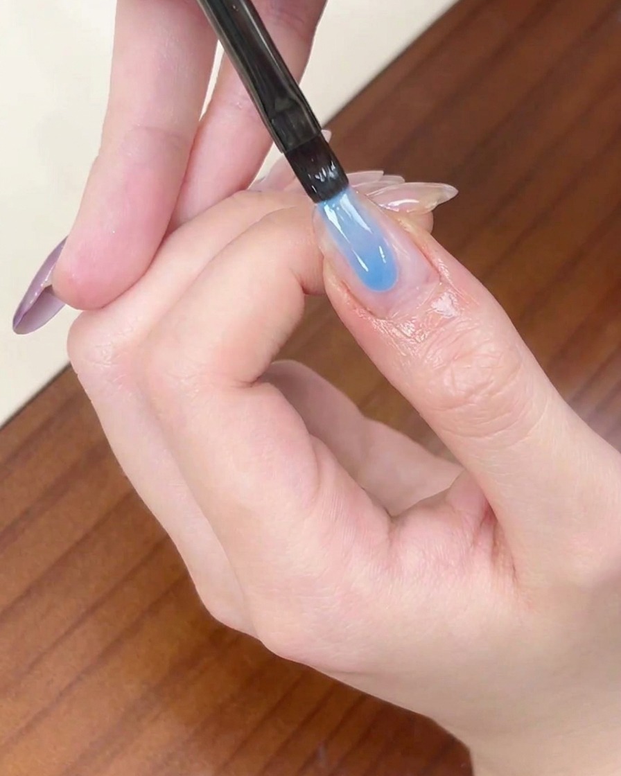 Un pinceau liner appliquant une bande de vernis gel bleu transparent sur le bord libre dun ongle en amande.