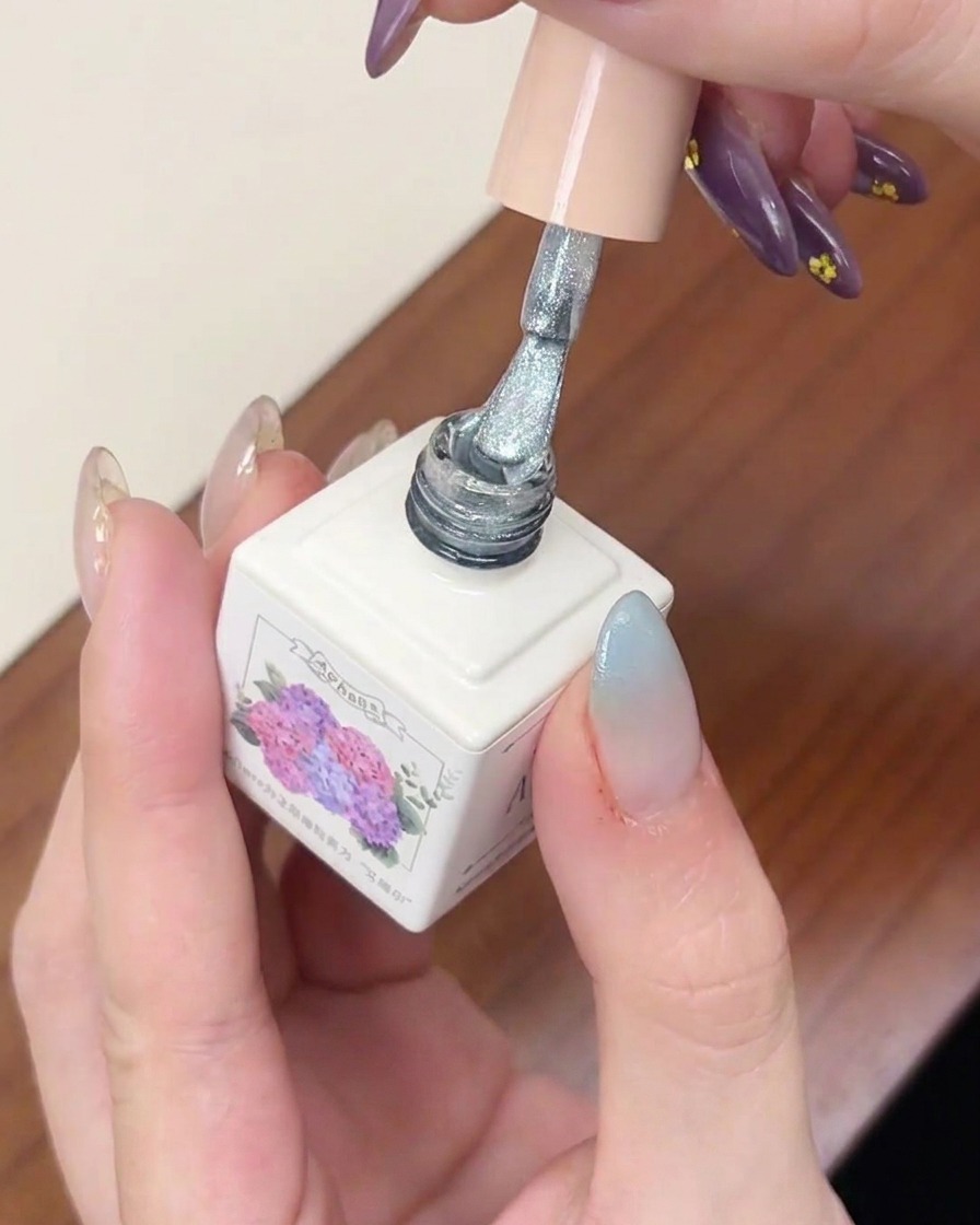 Un pinceau à vernis gel appliquant une couche de gel cat-eye argenté scintillant sur un ongle avec un dégradé bleu.