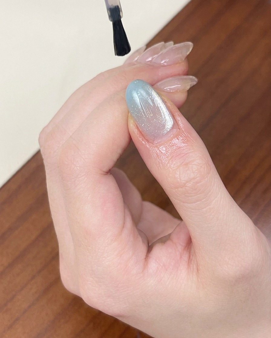 Un pinceau appliquant un top coat transparent brillant sur un ongle bleu clair effet œil-de-chat scintillant.