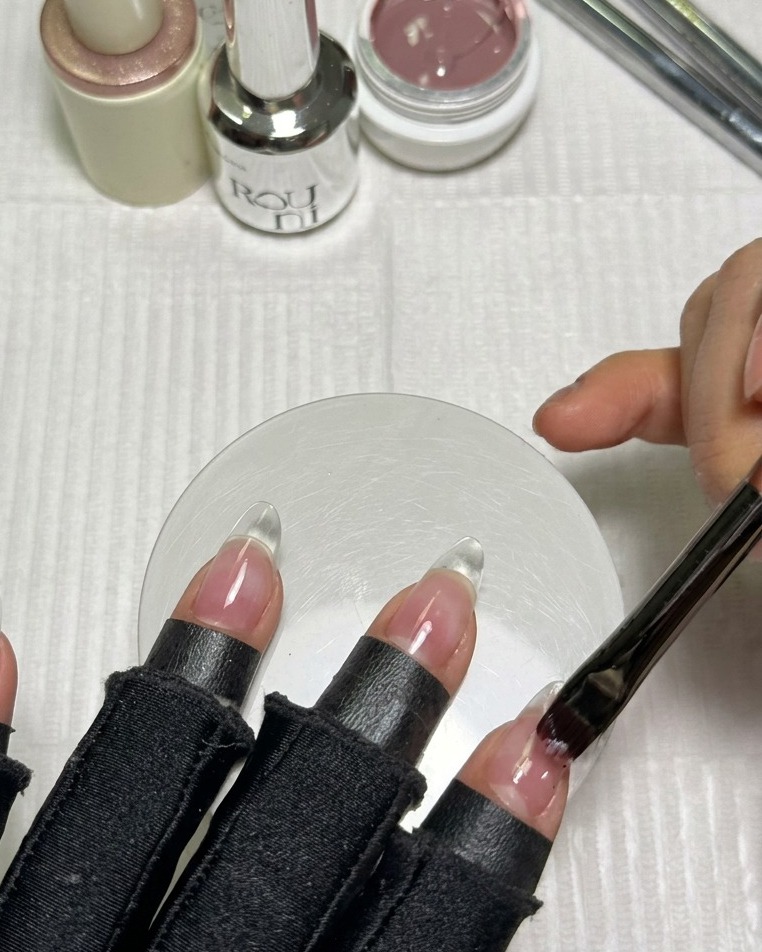 Un pinceau appliquant une fine couche de vernis gel nude rosé sur une extension dongle transparente en forme damande.