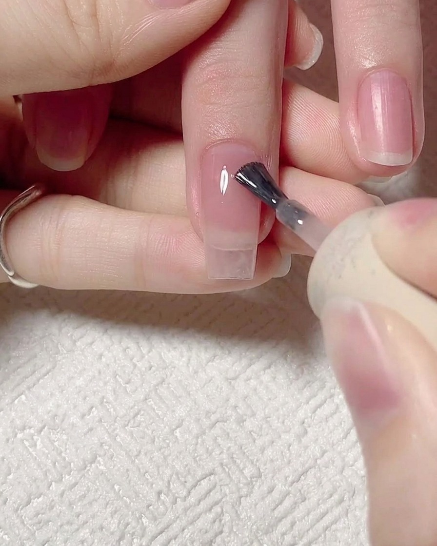 Un pinceau appliquant un gel rose jelly transparent sur un ongle naturel propre et préparé.