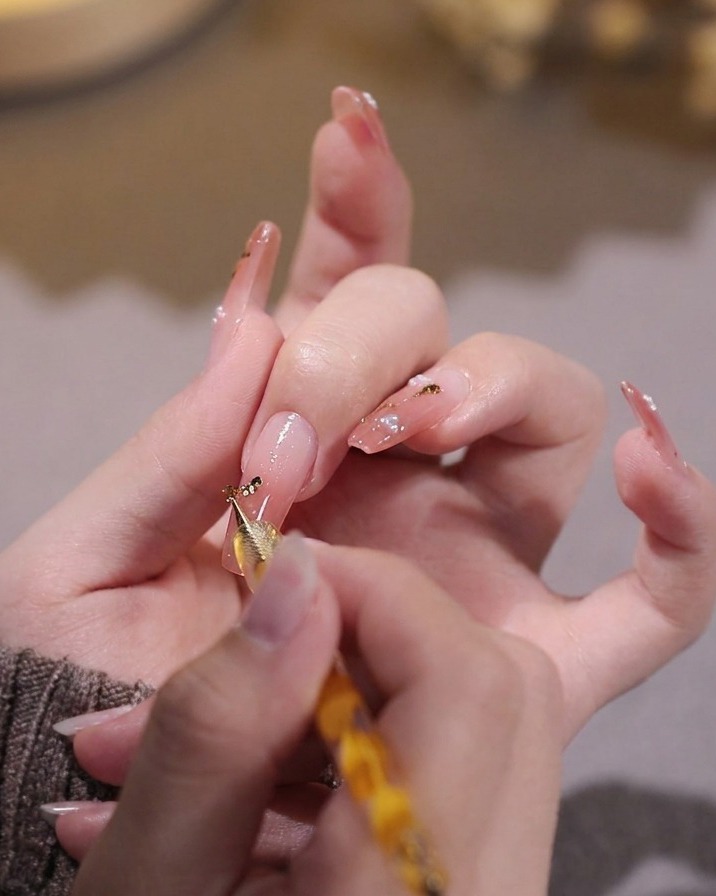 Une main utilisant un outil à points pour placer de minuscules flocons de feuille dor sur un ongle peint en gel rose.