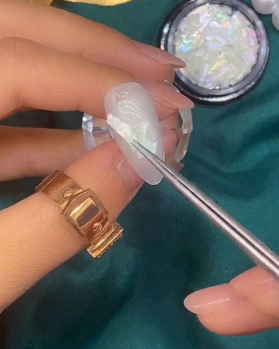 Un pinceau déposant de minuscules flocons de feuille dor sur un ongle déjà décoré de coquillages.