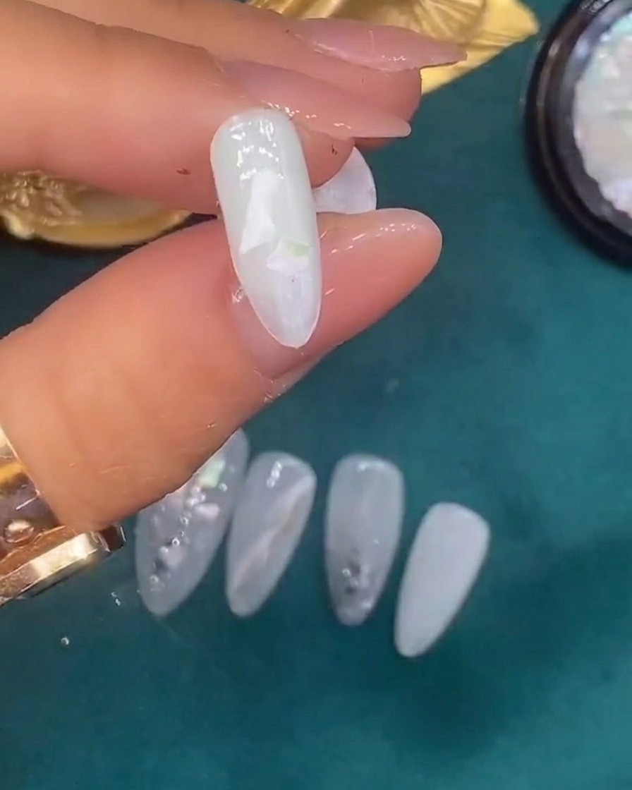 Une pince déposant des coquillages décoratifs sur un ongle recouvert de gel blanc translucide humide.