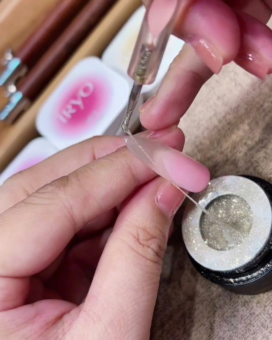 Un outil de dotting soulevant un long brin fin et élastique de spider gel à paillettes argentées dun pot.