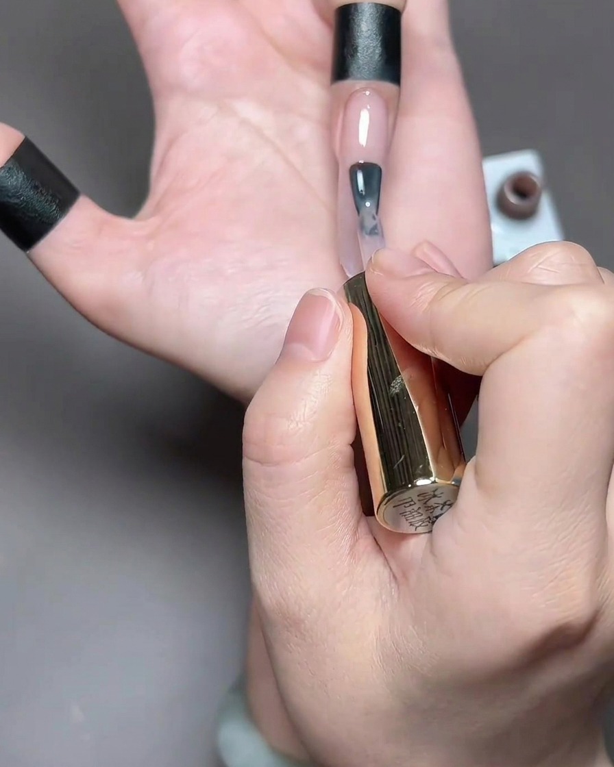 Un pinceau appliquant un vernis gel translucide aux tons marron clair sur un long ongle naturel.